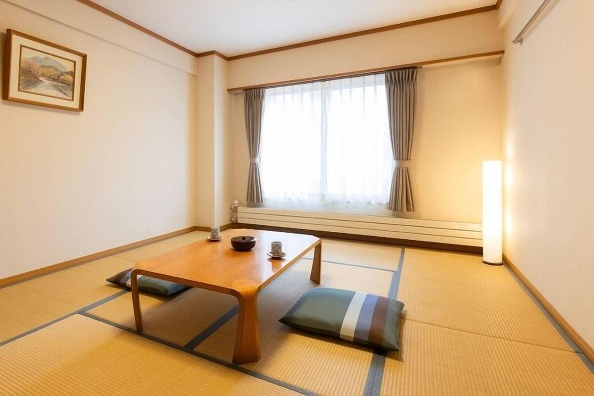 Una stanza tradizionale giapponese con pavimento in tatami, un tavolino in legno, un cuscino, un quadro appeso e una finestra con tende.