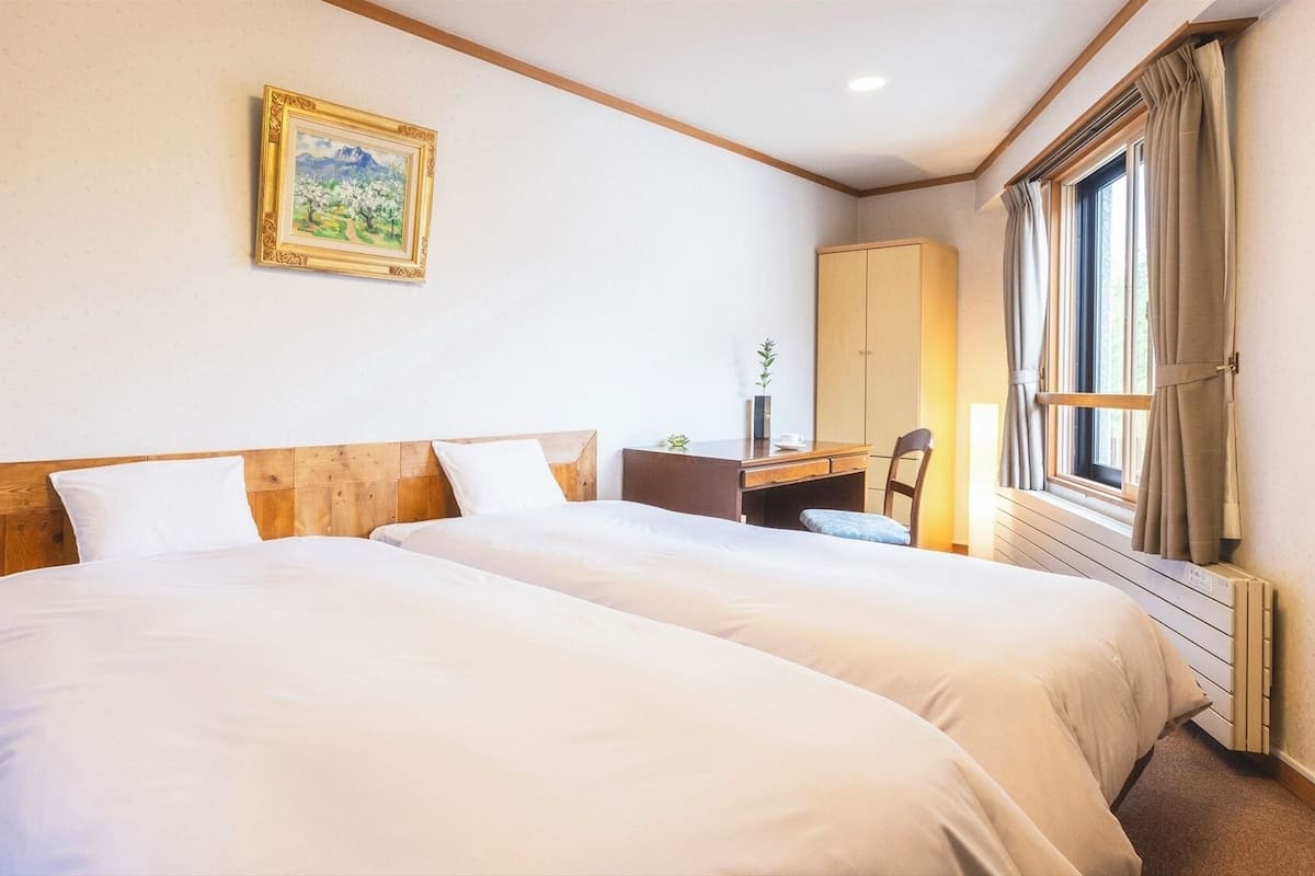 Camera d'albergo con due letti, una scrivania, una sedia, una finestra con tende e un quadro appeso al muro.