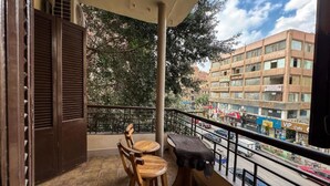 A balcony with wooden chairs and a table, overlooking a busy street with buildings and shops.