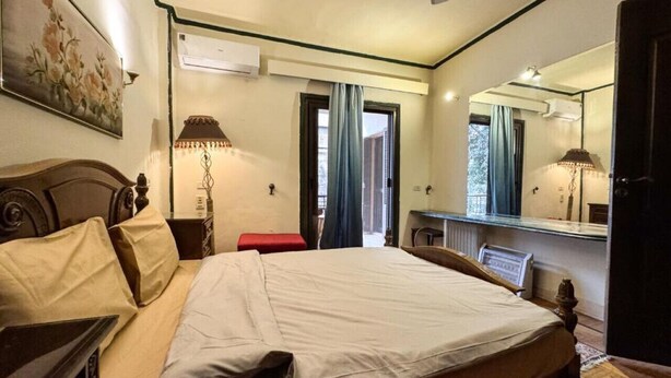 A hotel room with a large bed, a desk, a chair, a mirror, and an air conditioning unit.