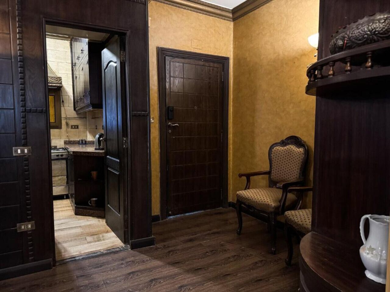 Una habitación con suelo de madera, una silla, una puerta y una cocina visible a través de una puerta abierta.