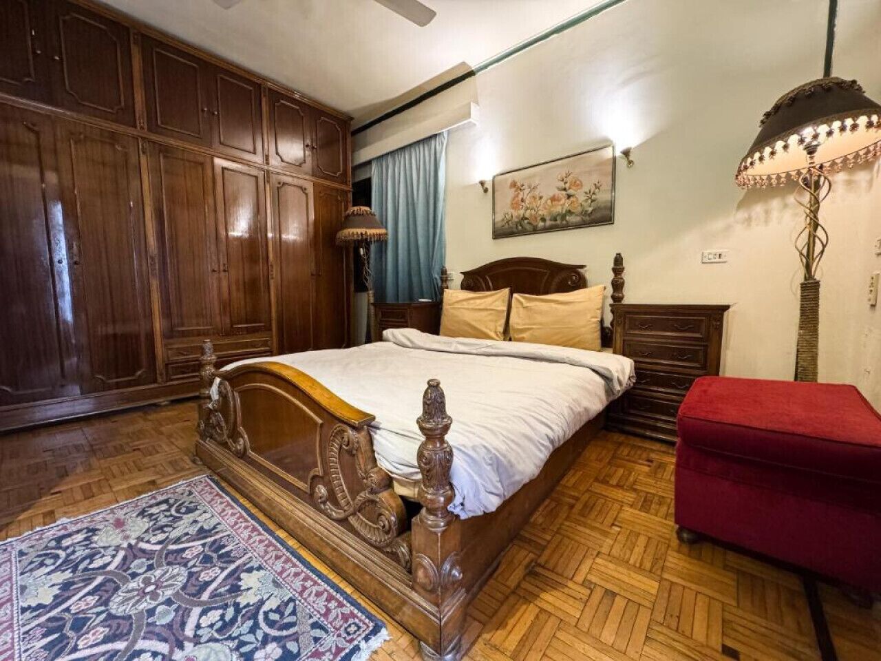 A bedroom with a wooden bed, a red ottoman, a wooden wardrobe, and a patterned rug.