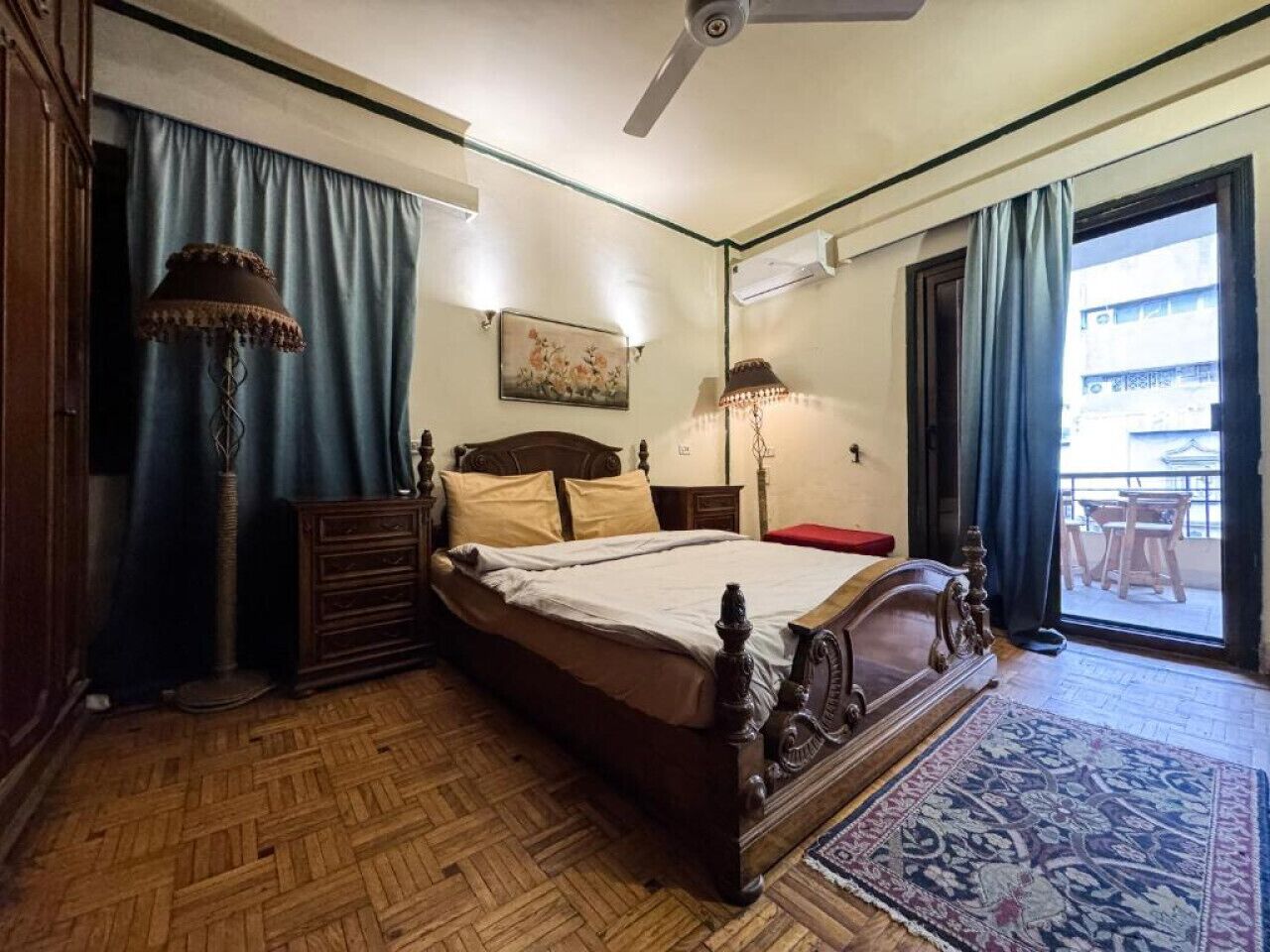 Una camera da letto con un letto in legno, una cassettiera, una lampada da terra e un'ampia finestra con tende.