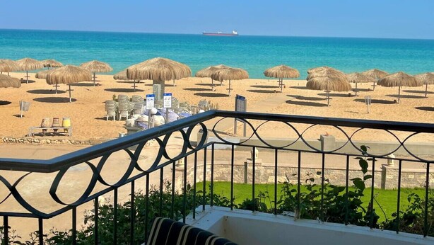 Vista a la playa desde un balcón con sombrillas de paja, sillas de descanso y un barco a lo lejos.