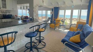 Amplia sala de estar con un ventanal grande con vistas al mar, una cocina con armarios blancos y una zona de bar con taburetes.
