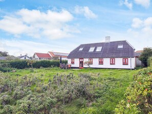 Una casa blanca con ventanas rojas y techo inclinado, rodeada de vegetación y otras viviendas.