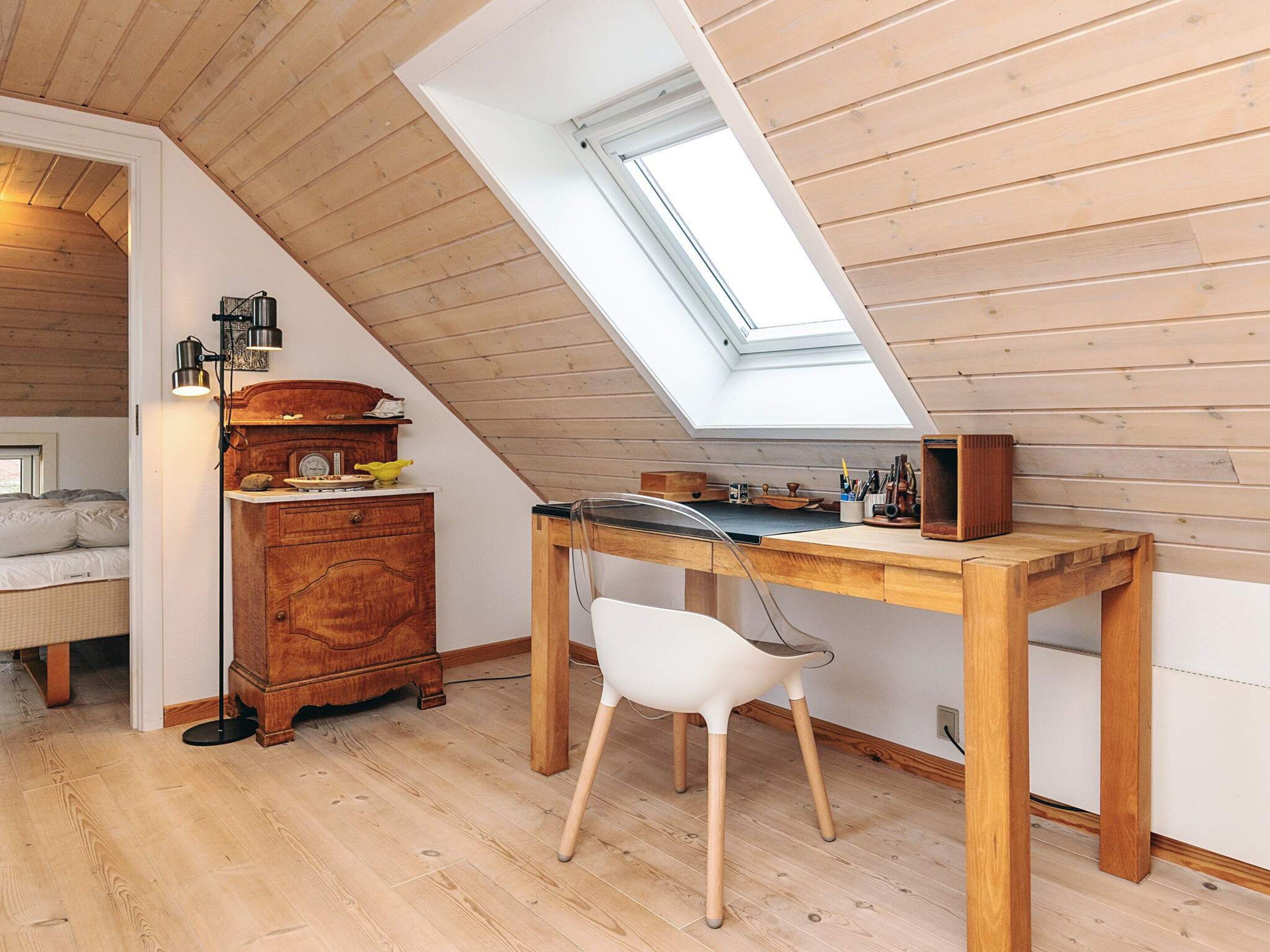 A cozy attic bedroom with a sloped ceiling, a skylight, a wooden desk, a white chair, and a vintage dresser.