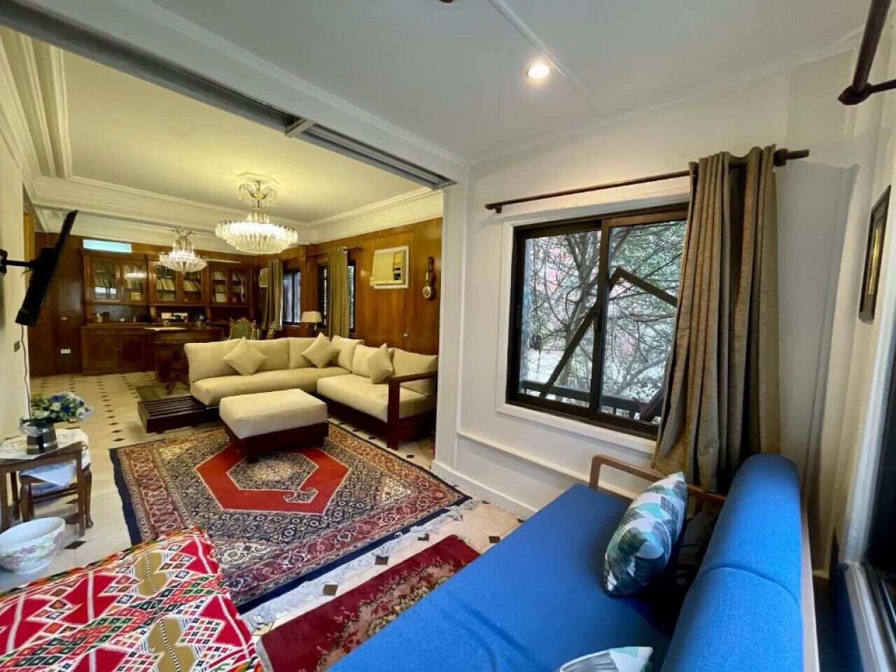 A living room with a blue sofa, beige armchairs, and a patterned rug. There is a large window with curtains, a chandelier, and a bookshelf in the background.