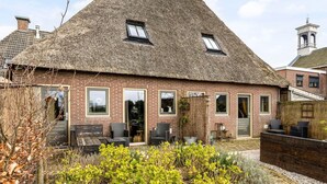 Une maison traditionnelle à toit de chaume, dotée d’une façade en briques, de nombreuses fenêtres et d’un petit jardin agrémenté d’un espace de détente extérieur.