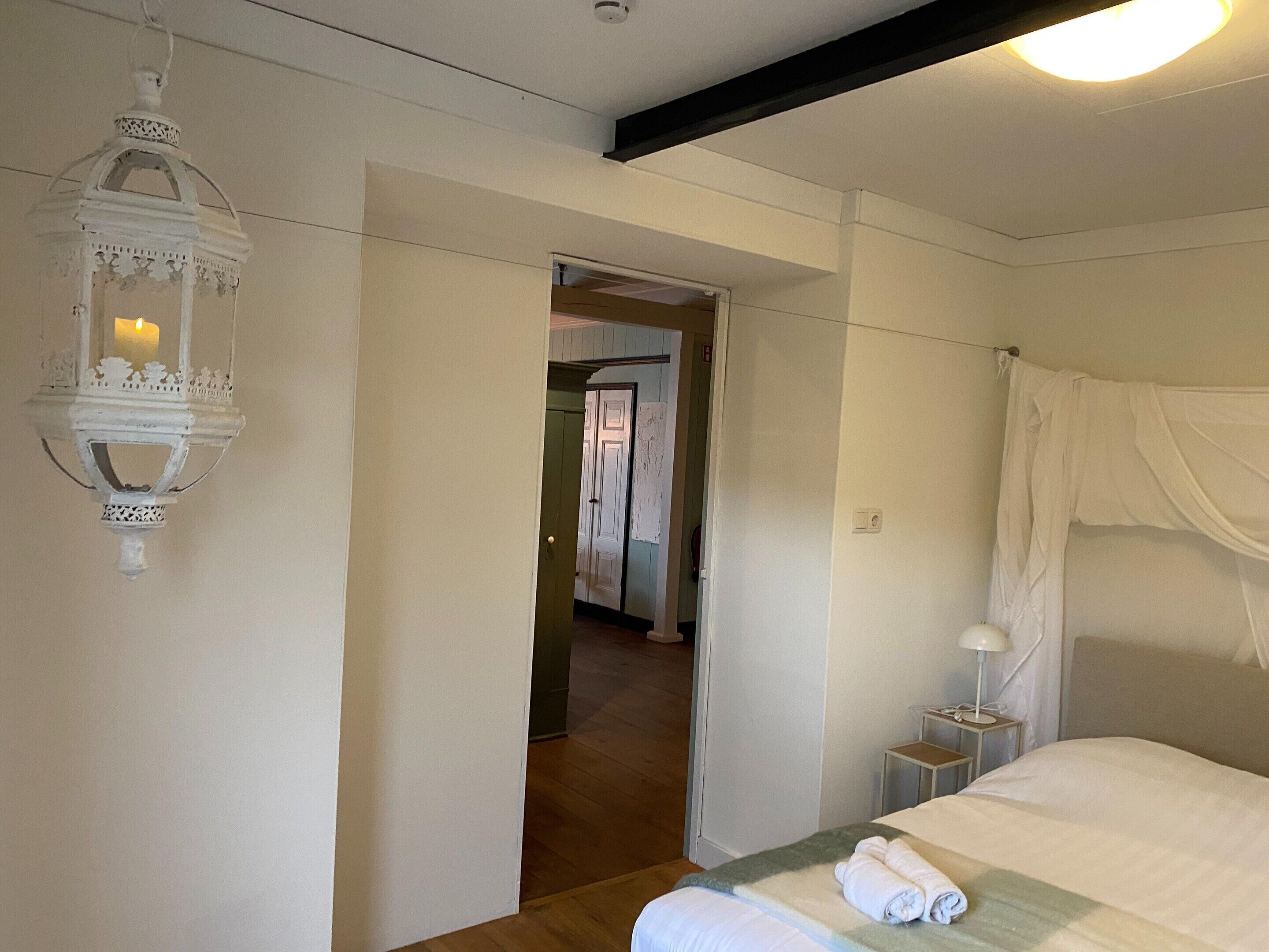 A bedroom with a bed, a nightstand, a lamp, a hanging lantern, and a door leading to another room.