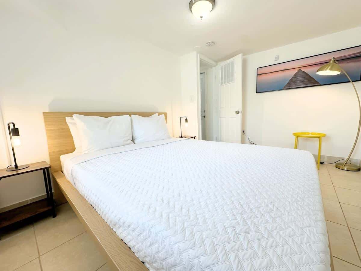 A neatly made bed in a room with a wooden headboard, a bedside table with a lamp, a yellow side table, and a painting on the wall.