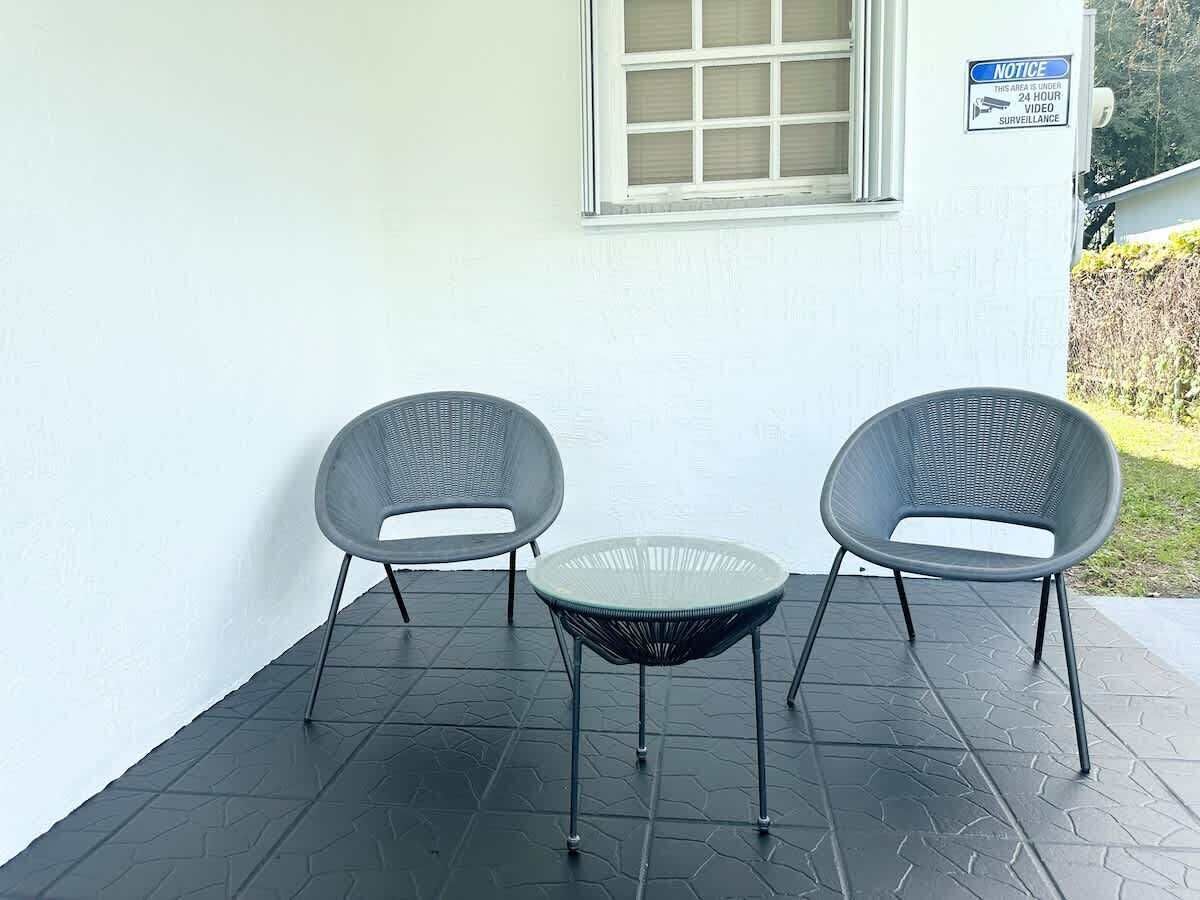 Un'area patio con due sedie e un tavolino, una finestra con persiane e un cartello che avvisa della videosorveglianza attiva 24 ore su 24.