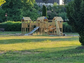 A playground with multiple wooden play structures, including a slide and climbing frames, set in a grassy area with trees and a building in the background. - Reihenhaus am Gross Labenzer See in Klein Labenz (Warin)