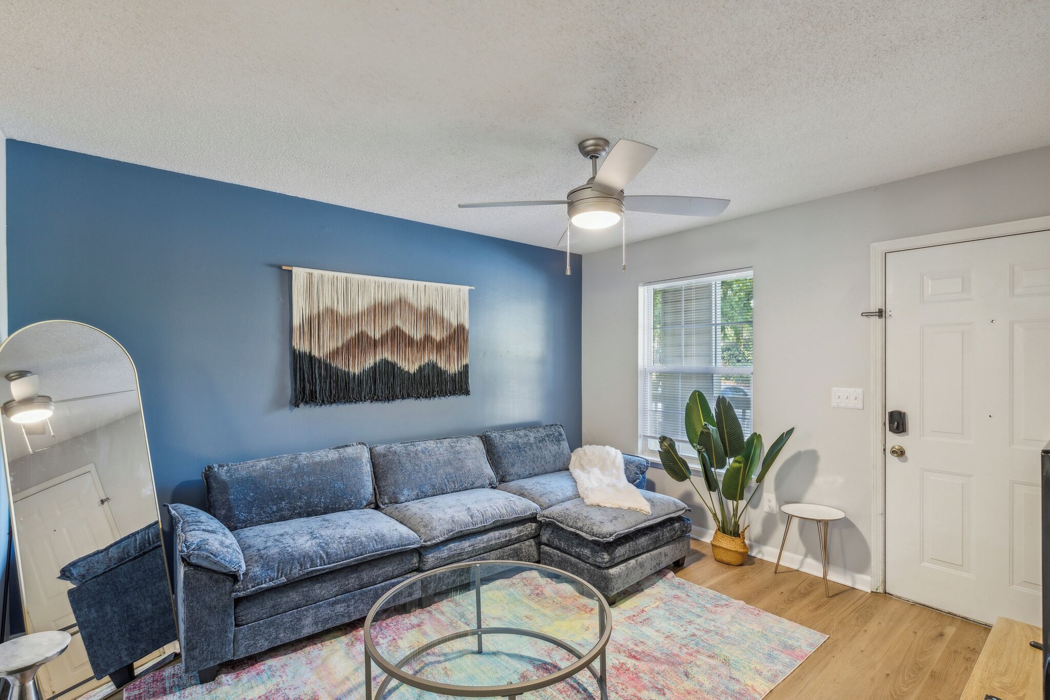 A living room with a blue wall, a sectional sofa, a ceiling fan, a large mirror, and a colorful rug.