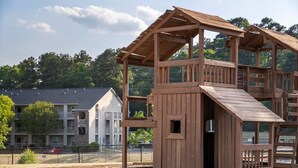 A wooden cabin structure with a sloped roof and a small door, surrounded by a fence and trees.