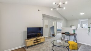 A modern living room with a flat-screen TV, a glass coffee table, and a yellow ottoman.