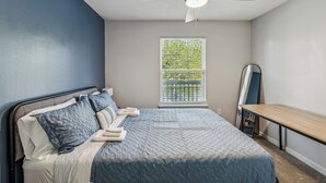 Une chambre avec un lit, un ventilateur de plafond, une fenêtre avec des stores et un bureau.
