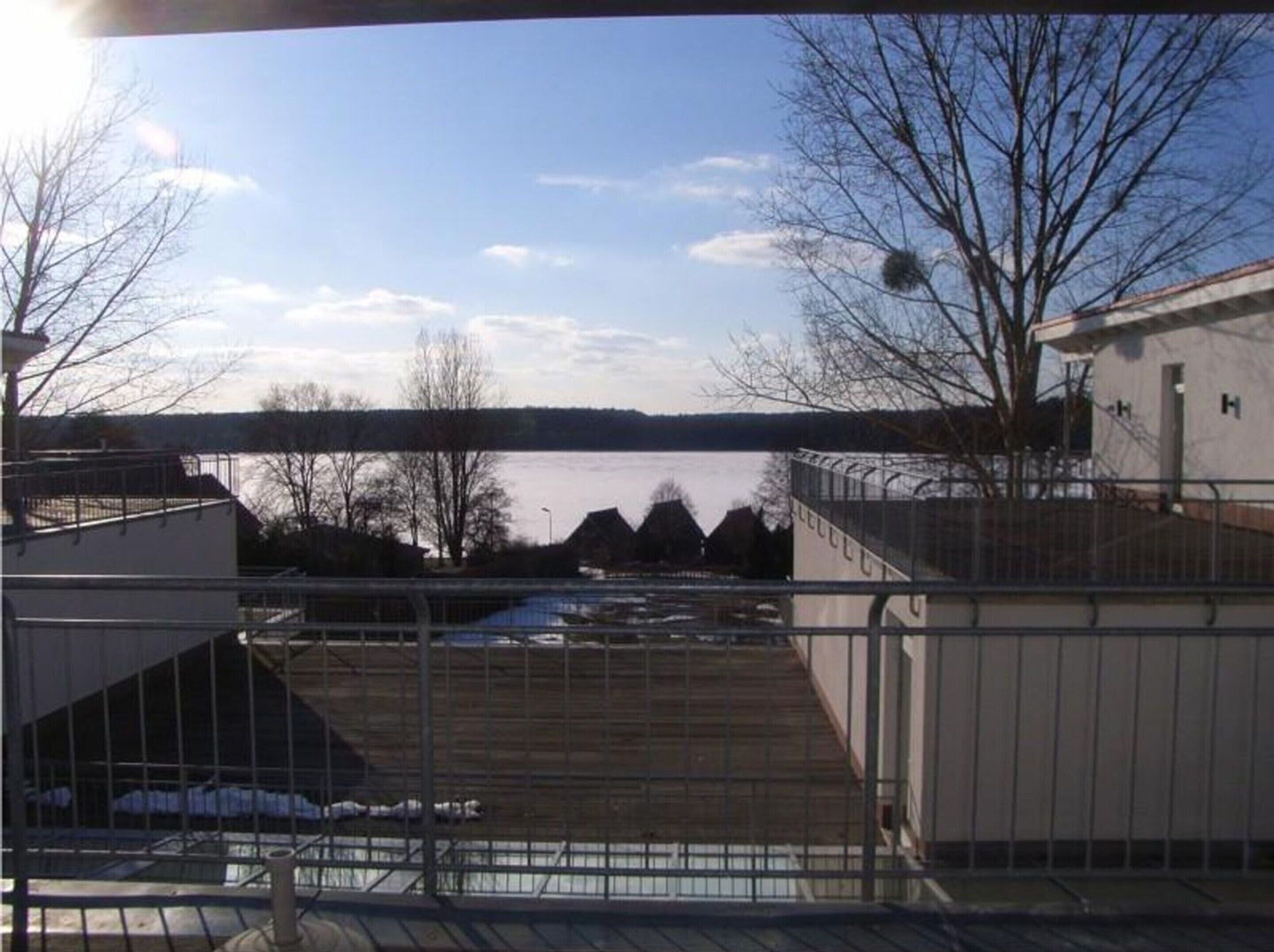 A view from a balcony overlooking a body of water with a distant shoreline, a building with a white exterior, and bare trees.
