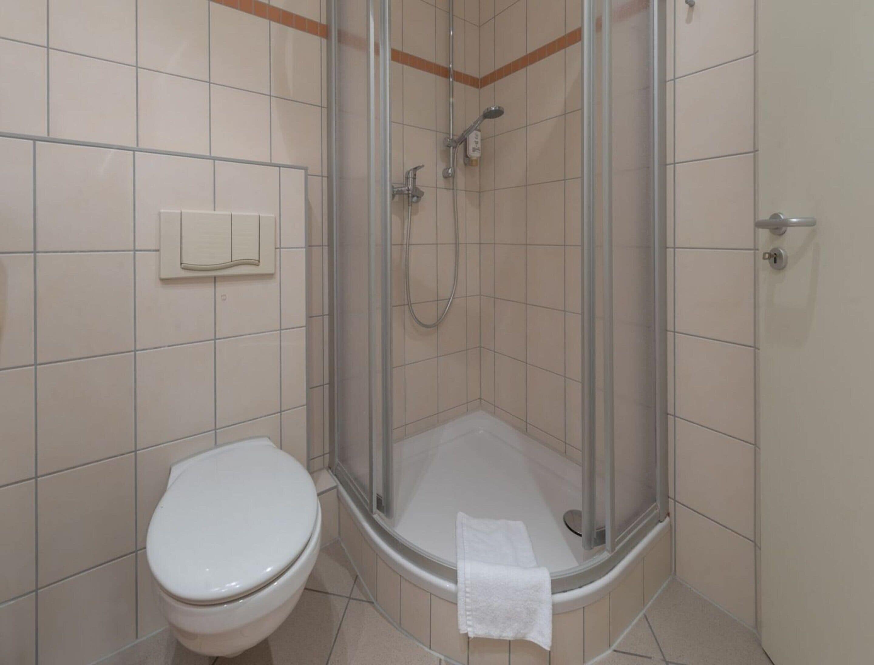 En lille, lukket brusekabine med glasdør, et toilet og et håndklæde på gulvet.