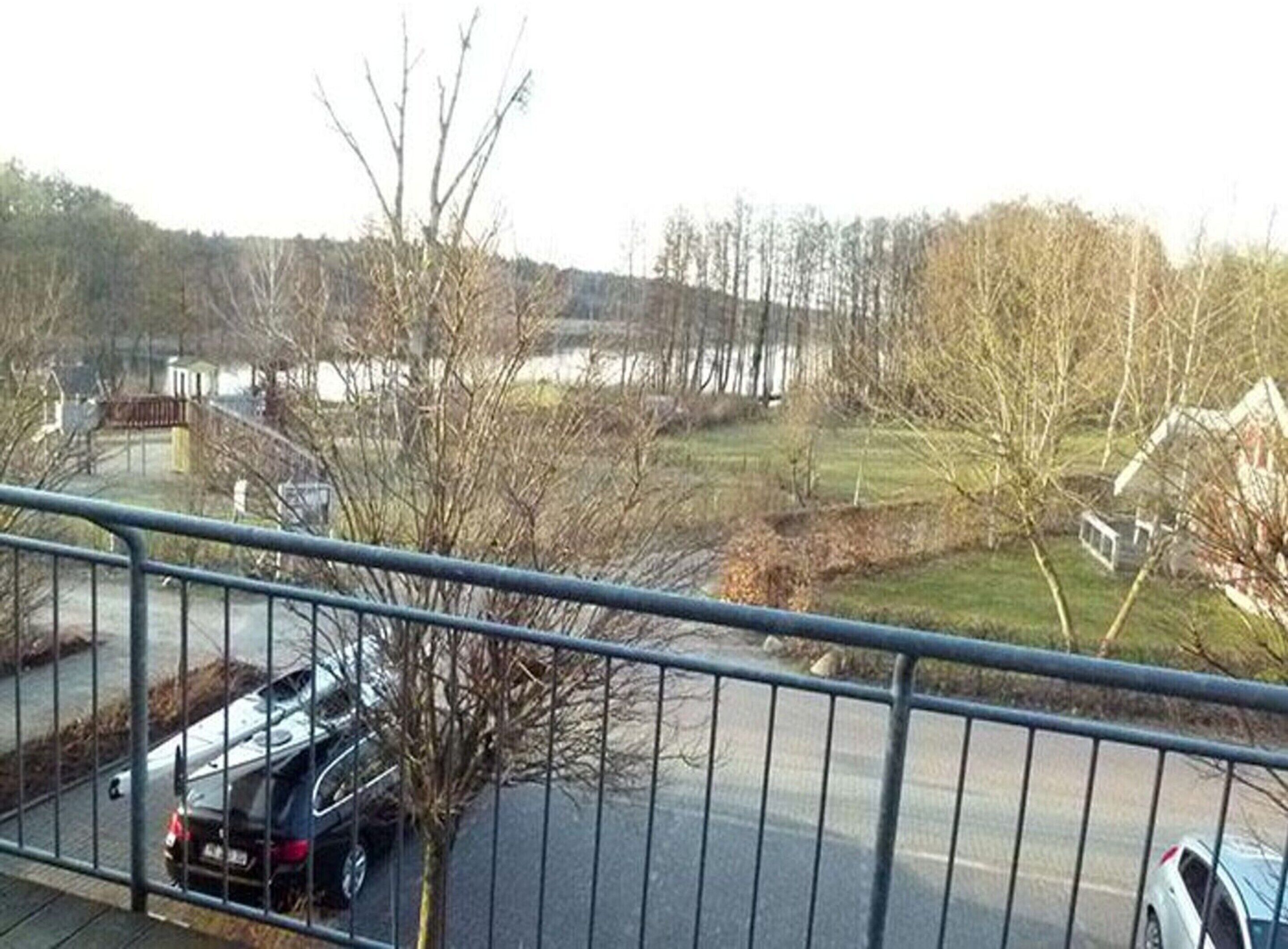 A view from a balcony overlooking a road with parked cars, a grassy area, and a body of water in the distance.