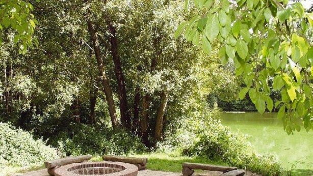 A circular stone fire pit surrounded by a brick border, set in a grassy area with benches and a pond in the background.