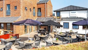 An outdoor seating area with multiple tables and chairs, umbrellas, and a brick building with a restaurant sign. - Apartment in Holiday Park in Vederso Klit (Ulfborg)
