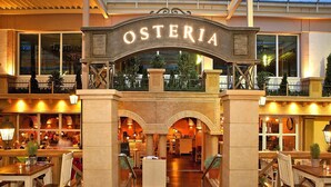 A restaurant named "OSTERIA" with a central archway, multiple dining tables, chairs, and a view of the outdoor area through large windows. - Appartements im Freizeitpark Weissenhauser Strand (Wangels)