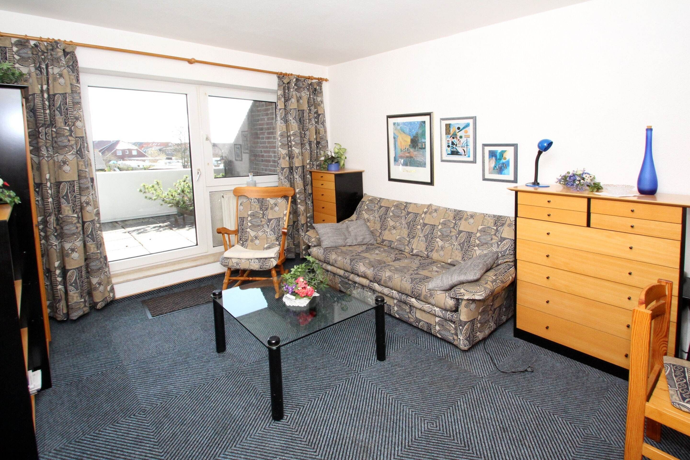 A living room with a patterned sofa, a glass coffee table, a wooden dresser, and a chair. There are framed pictures on the wall and a plant on the dresser.