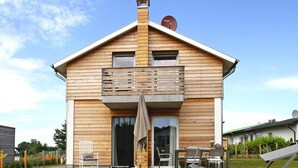 Ein zweistöckiges Holzhaus mit Balkon, Schiebeglastür und Terrasse mit Gartenmöbeln.