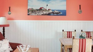 Une pièce avec un mur recouvert de panneaux blancs, un tableau représentant un phare, une table sur laquelle se trouve un saladier en verre et des chaises à rayures.