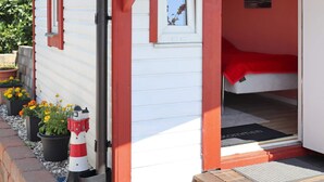 Un petit bâtiment d’un seul étage, à l’extérieur rouge et blanc, avec une porte bien visible et un petit porche agrémenté de plantes en pot.