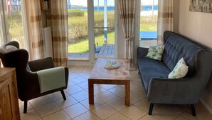 Ein Wohnzimmer mit einer Couch, einem Sessel und einem kleinen Tisch. Große Fenster mit Vorhängen bieten Blick auf einen Garten und ein Gewässer.