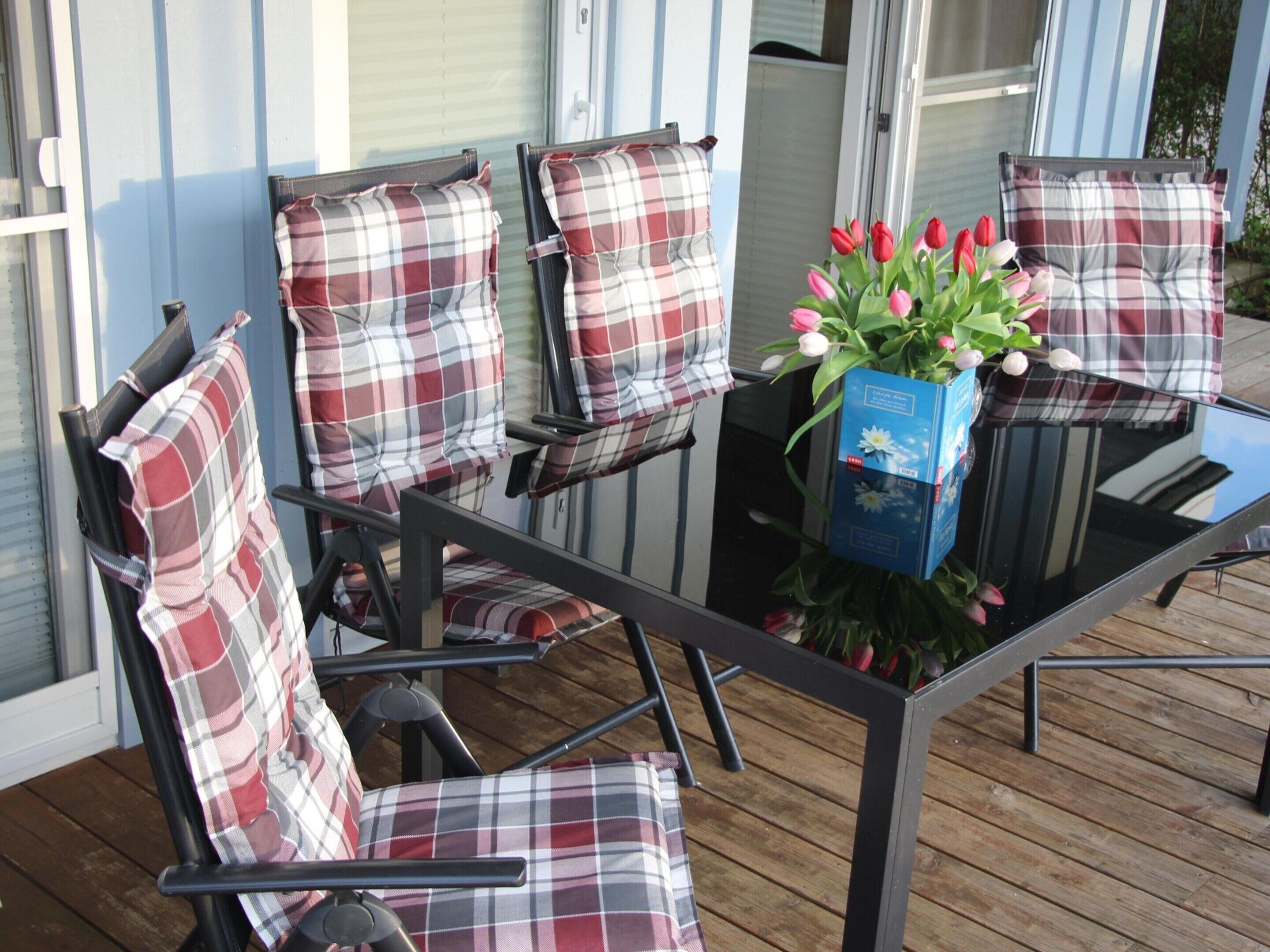 A glass-top table with a vase of flowers and plaid-patterned chairs on a wooden deck.