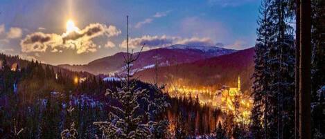 A snowy landscape with a village illuminated by lights, surrounded by mountains and trees.