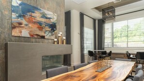 A modern dining area with a long wooden table, grey upholstered chairs, and a large abstract painting above the fireplace.