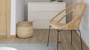 Una habitación minimalista con una puerta de madera, un aparador blanco, una silla de mimbre y un espejo grande en la pared.