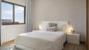 Un dormitorio con una cama grande, una mesita de noche, una lámpara y vista al mar por una ventana.