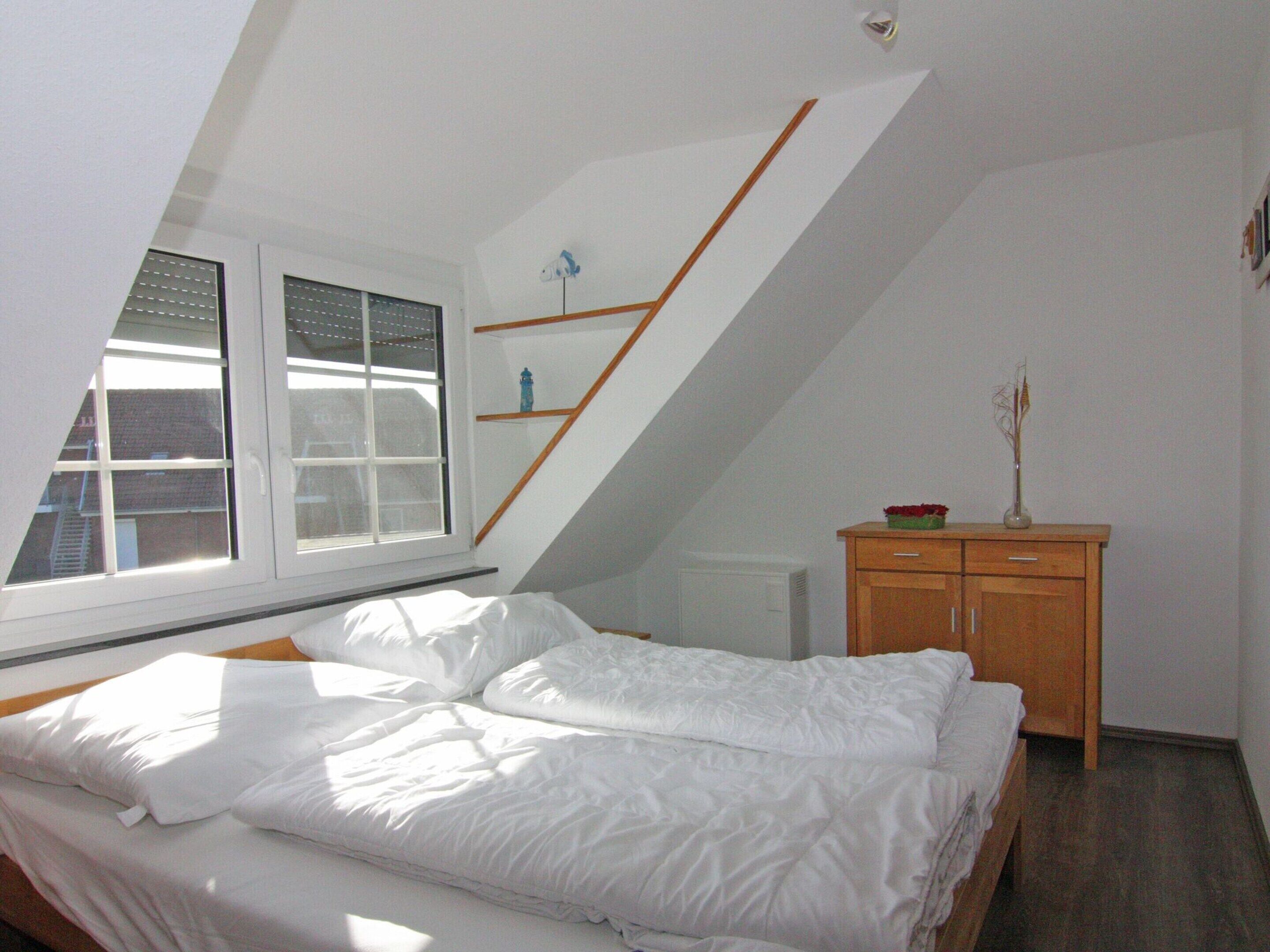 A bedroom with a sloped ceiling, a bed with white bedding, a wooden dresser, and a window with blinds.