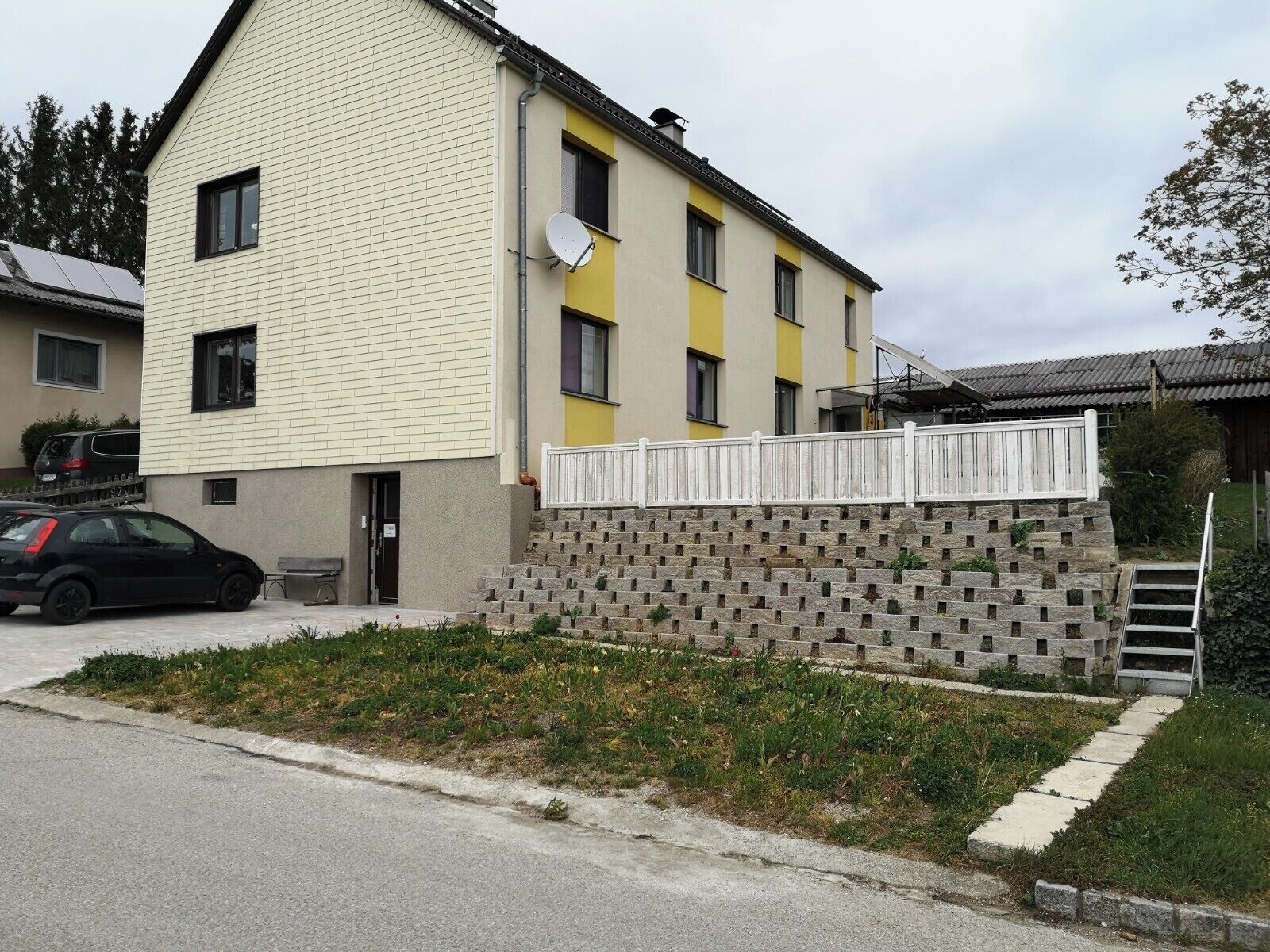 Ein zweistöckiges Gebäude mit gelb-beiger Fassade, einer Satellitenschüssel und einem kleinen Gartenbereich mit einer Steinmauer.