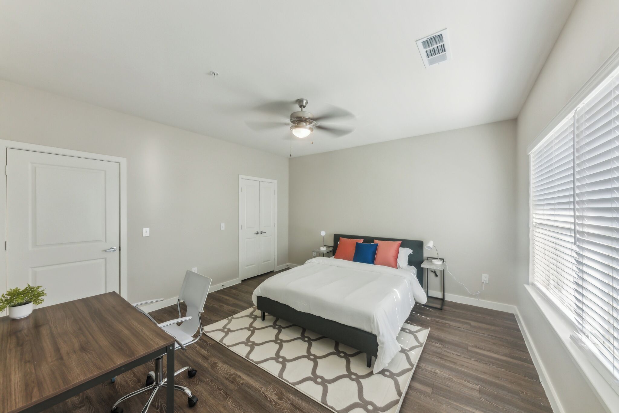 A bedroom with a bed, a desk, a chair, a ceiling fan, and a window with blinds.