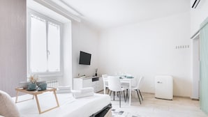 Una habitación moderna y bien iluminada con una cama, una mesa de comedor con sillas, un televisor montado en la pared y un refrigerador pequeño.