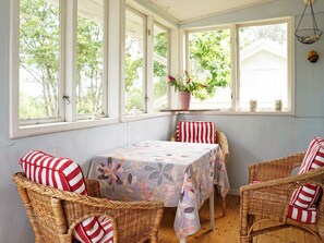 Una habitación con una mesa cubierta con un mantel floral, sillas de mimbre con cojines a rayas y una ventana con vistas a árboles.
