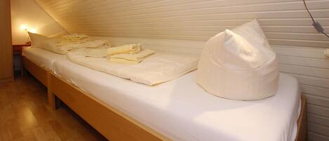 A single bed with neatly folded towels and a pillow, a bedside lamp, and a power outlet on a wooden floor.