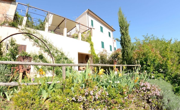 Un bâtiment blanc avec des volets verts, un balcon et un jardin où poussent différentes plantes et fleurs.