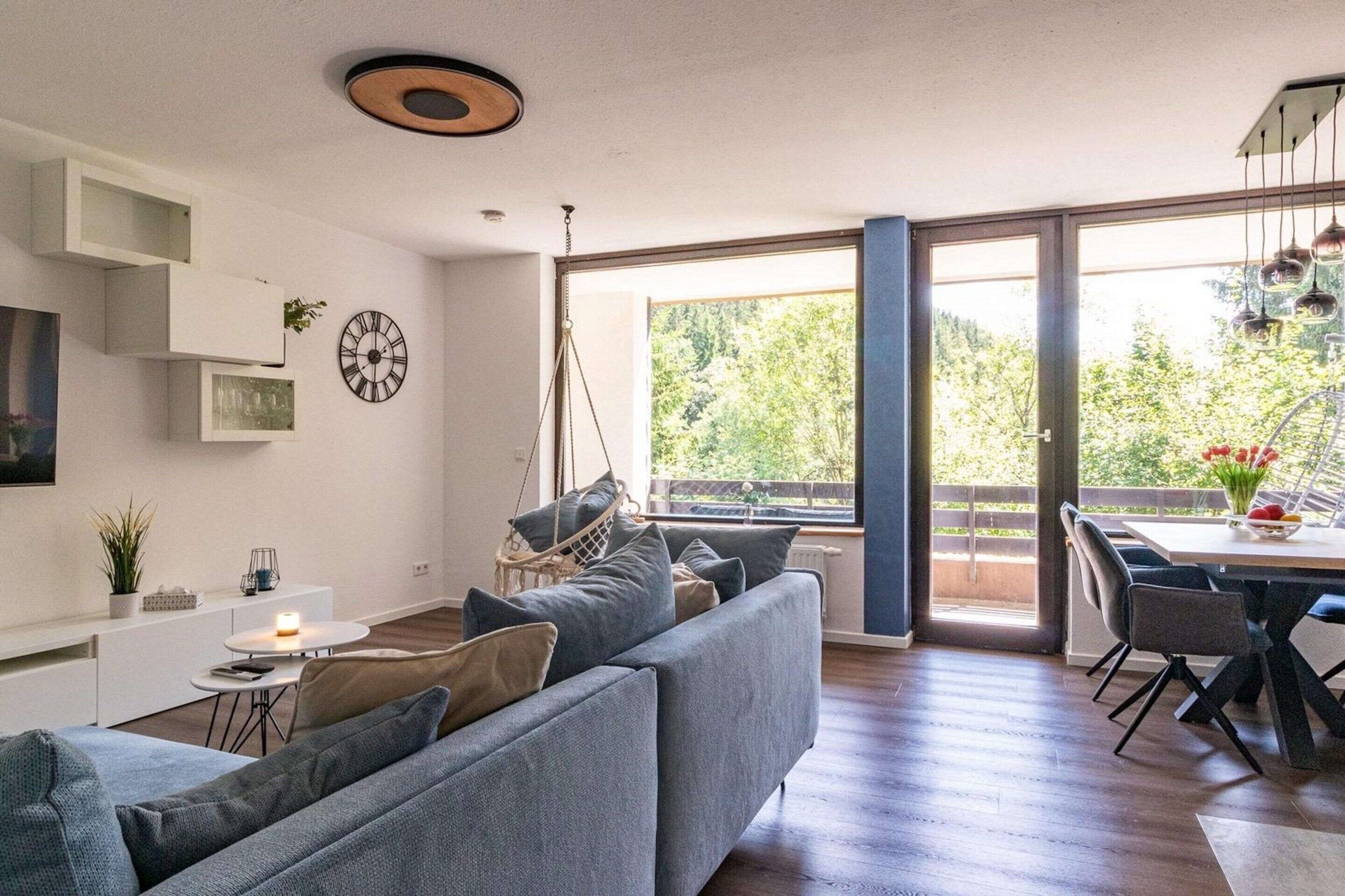 A modern living room with a grey sofa, a dining table with chairs, a hanging light fixture, and a large window with a view of greenery.