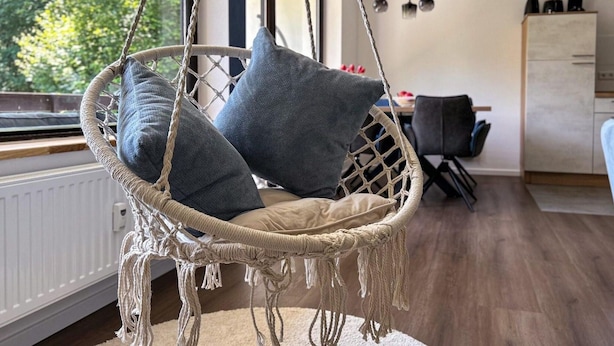 Una habitación con una silla colgante de macramé y cojines, un radiador blanco, suelo de madera, una zona de comedor con mesa y sillas, y una cocina con armarios y electrodomésticos.
