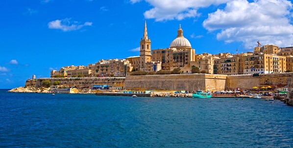 Un paisaje urbano costero con edificios históricos, una iglesia destacada con cúpula y campanario, y un puerto con barcos.