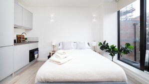 Ein modernes Schlafzimmer mit einem großen Bett, einer kleinen Küchenzeile und Blick auf ein Ziegelgebäude durch ein Fenster.