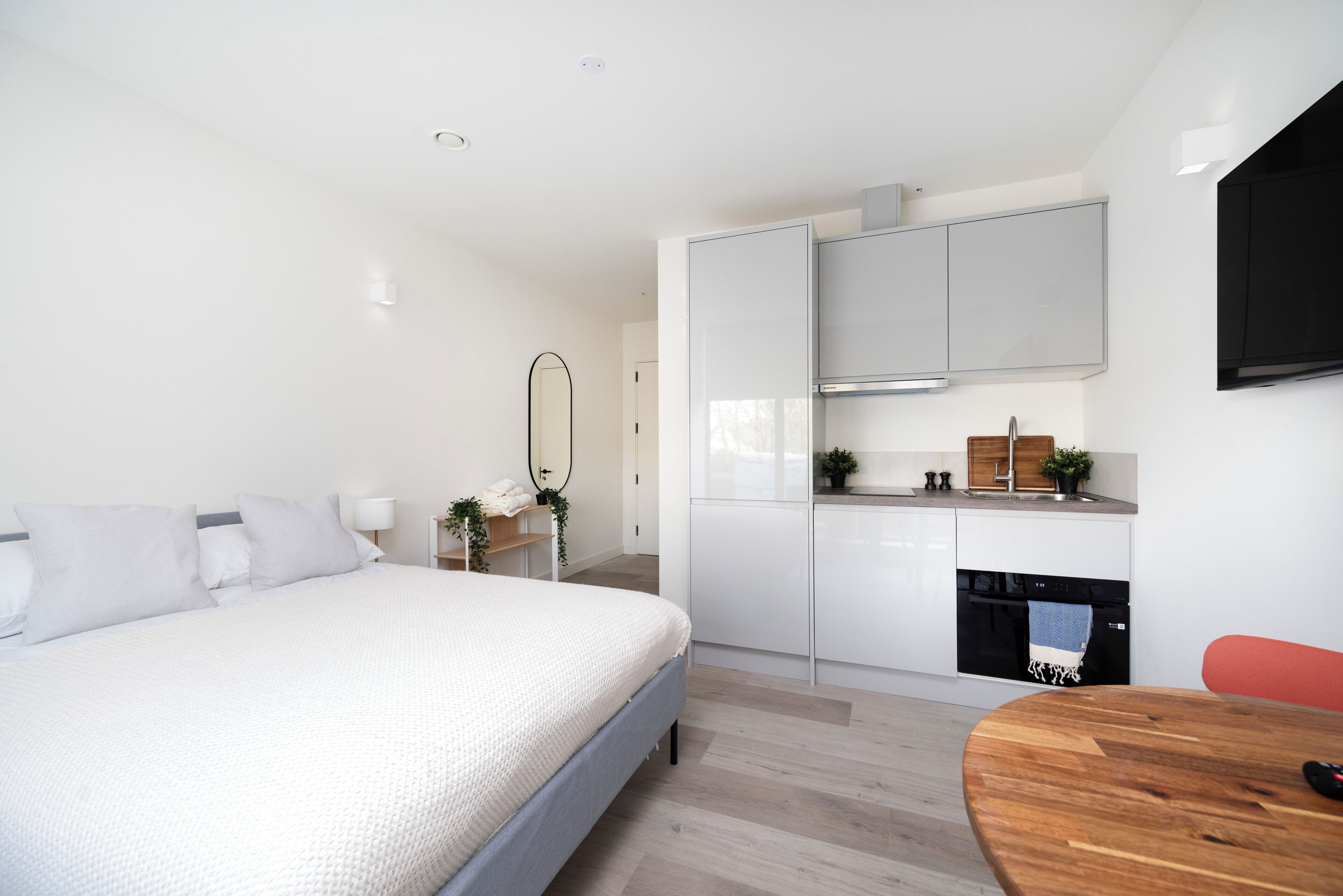Ein modernes Schlafzimmer mit einem großen Bett, einem Holztisch und einer Küchenzeile mit weißen Schränken und einem schwarzen Backofen.