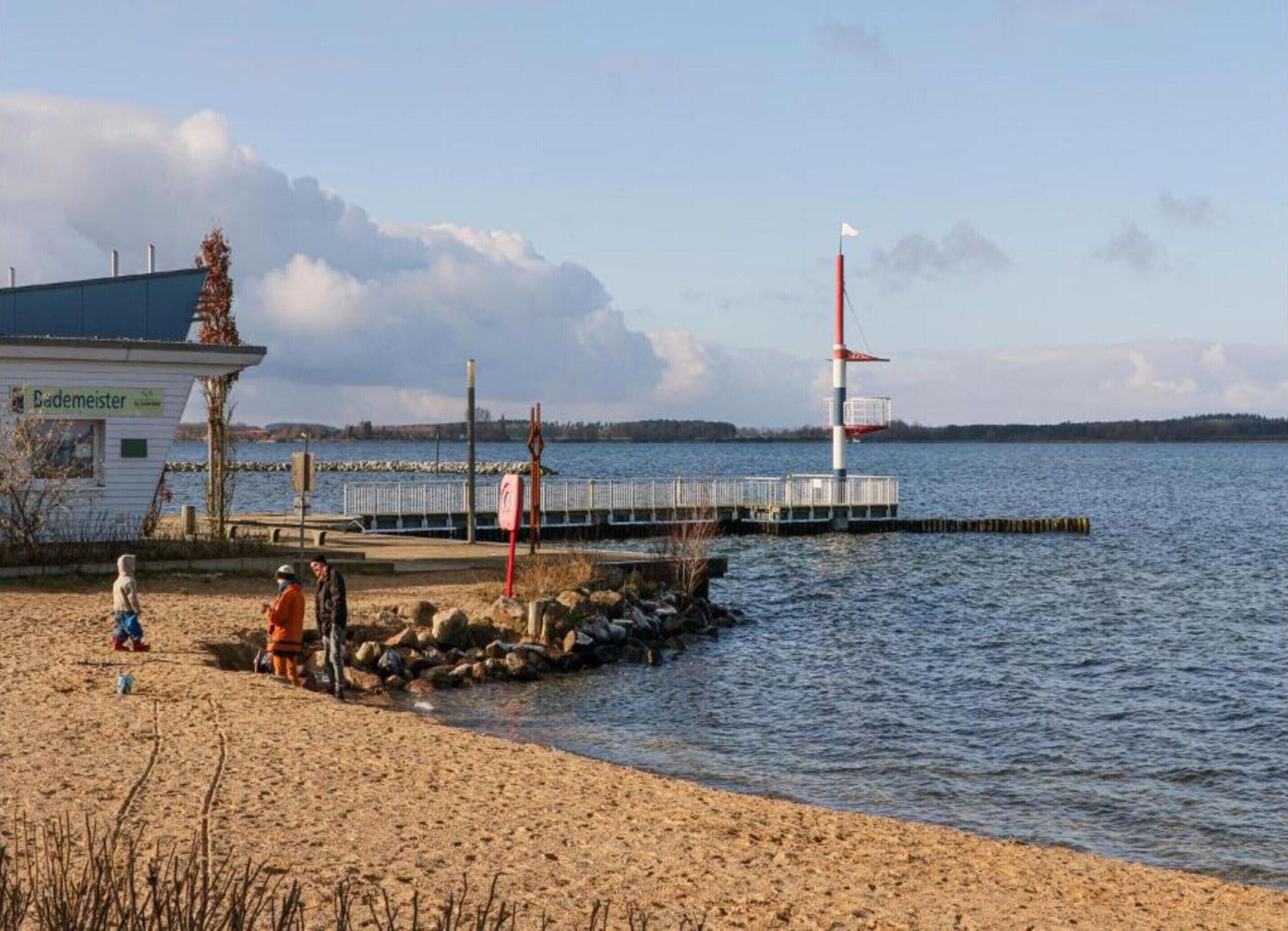 A beach with a lighthouse, a small building labeled 'Bademeister', and a rocky shoreline.